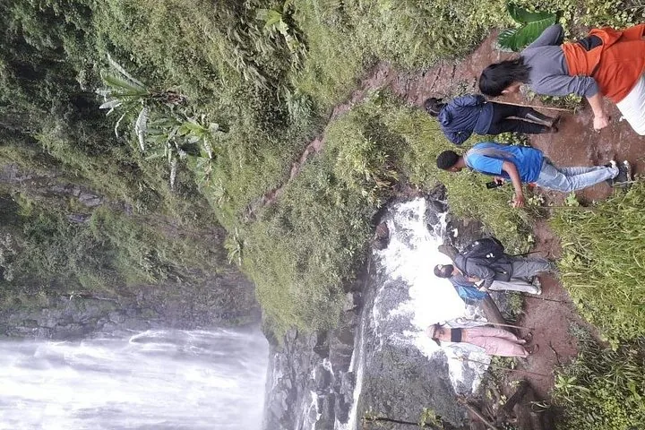 A group of people hike along a muddy trail near a waterfall.